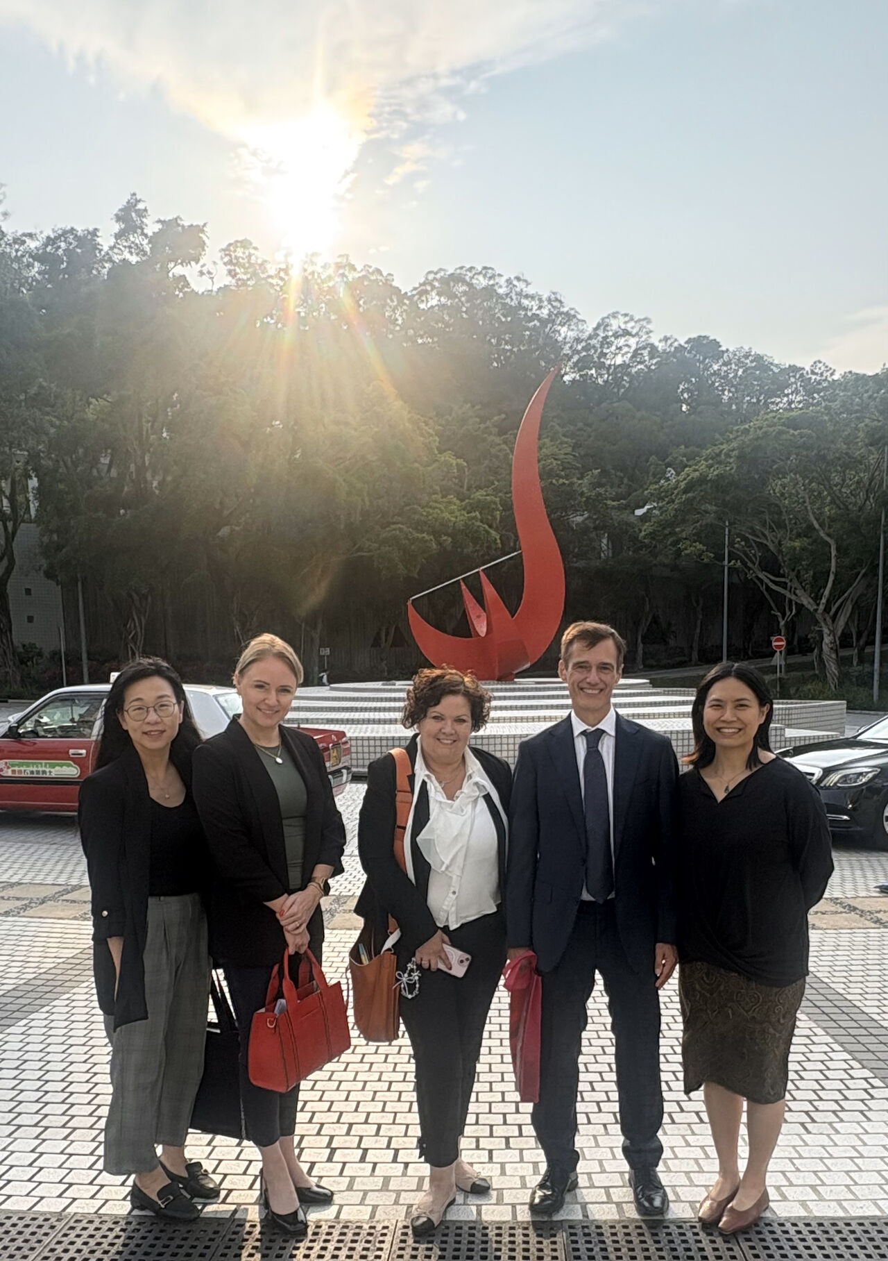 A group photo featuring Macquarie University Pro Vice-Chancellor (Graduate Research Prof. Simon HANDLEY (second right), Dr. Louise DWYER (second left), Operations Director of Graduate Research, and Hedda PAISLEY (center), Executive Director of Philanthropy and Alumni Relations, alongside the HKUST team, taken in front of the iconic "Red Bird" sculpture at the Piazza.